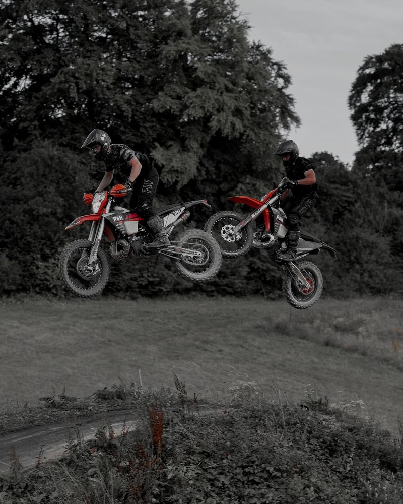Two riders side by side in the air over a jump on our track, with woodland behind