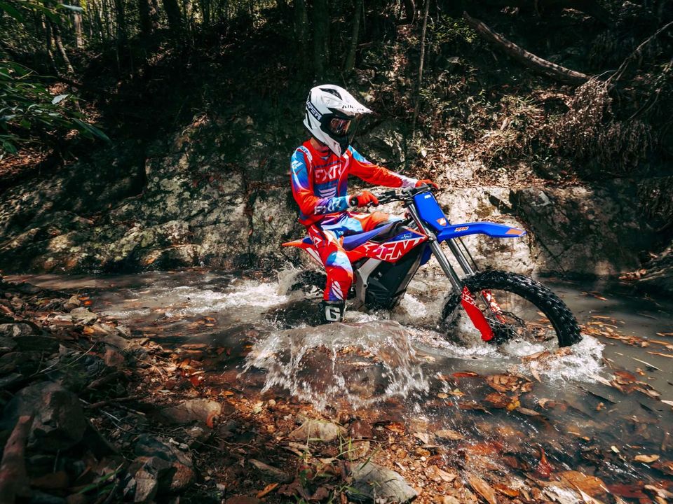 A rider in red and blue RFN kit splashing an RFN electric bike through a woodland stream